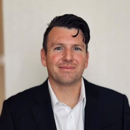 Headshot of Michael Rime, the finalist for Montezuma County administrator, in a dark jacket and white shirt against a pale wall.