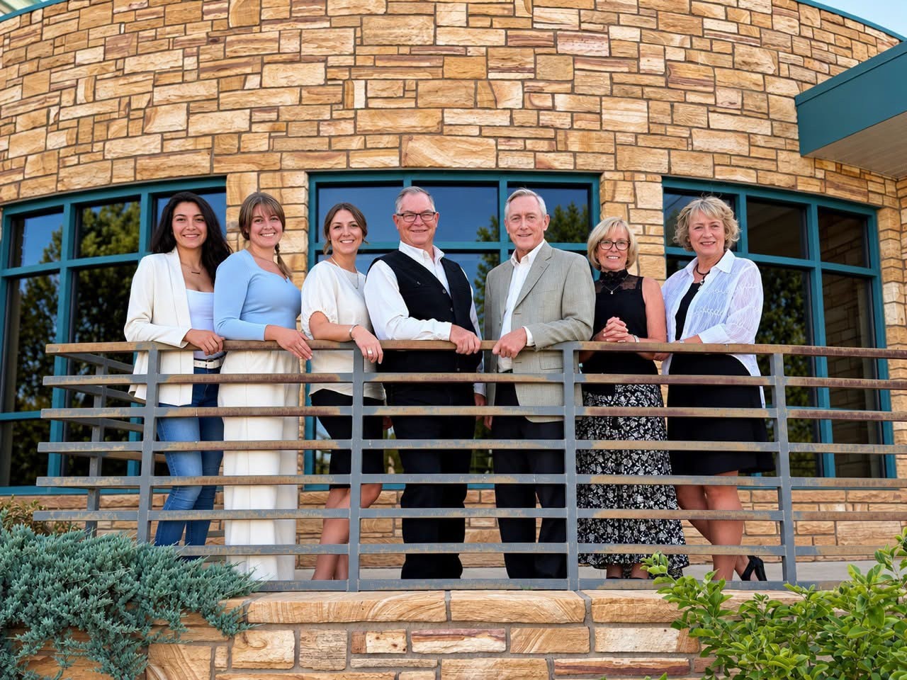 Seven members of the Cortez City Council stand in a row behind a metal railing in front of a curved sandstone building with teal-framed windows: five women and two men, dressed in a mix of white, light blue, black and gray.