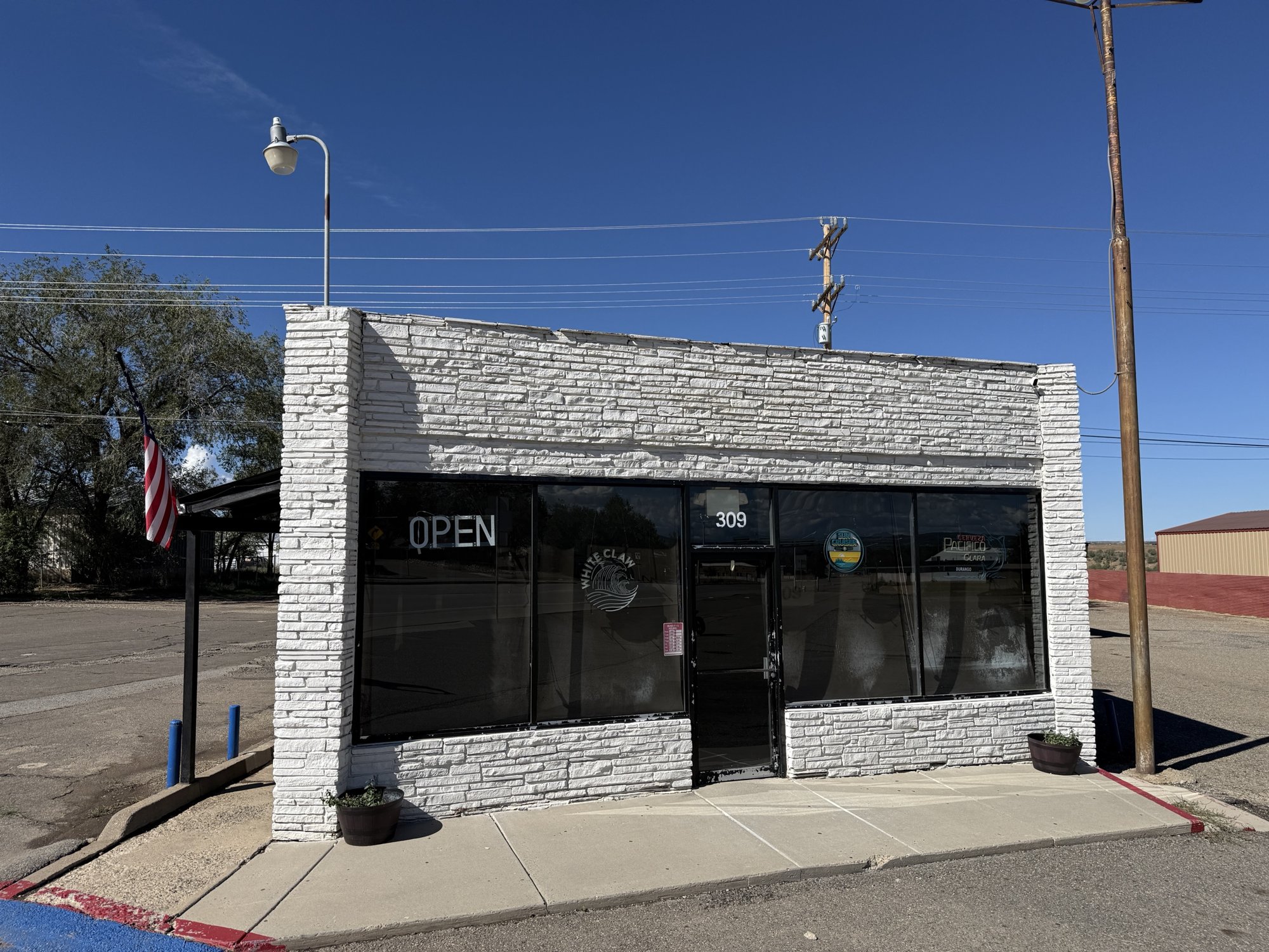The one-story building at 309 N. Broadway in Cortez, with a white stone front, large tinted windows, an Open sign, beer signs in the glass and an American flag on the side, under a clear blue sky.
