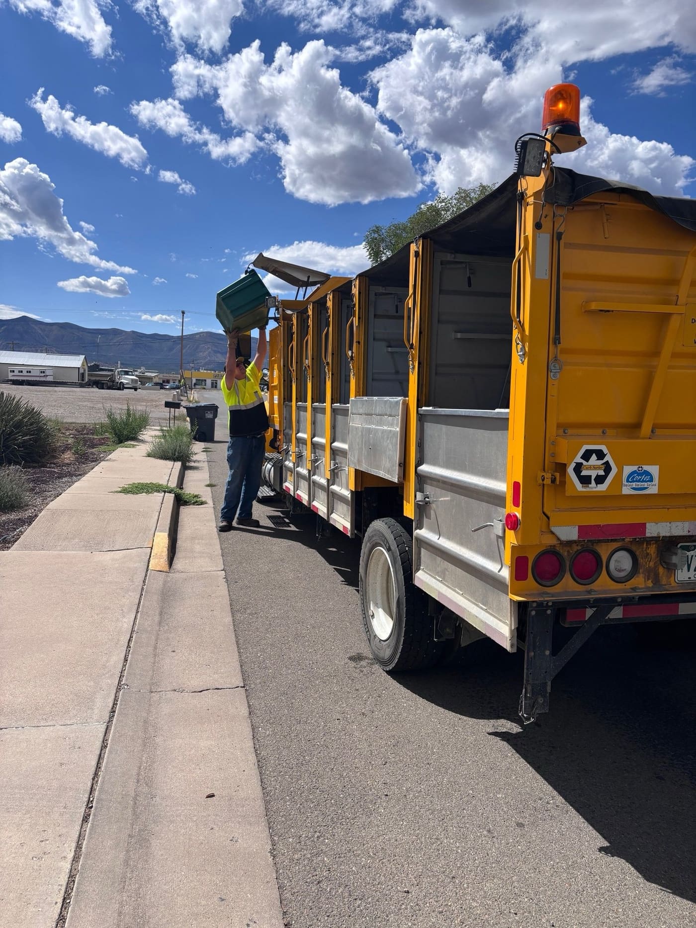 A worker in a yellow safety shirt lifts a green recycling bin over his head and empties it into a yellow City of Cortez curbside recycling truck parked along a curb, with mountains and clouds in the background.