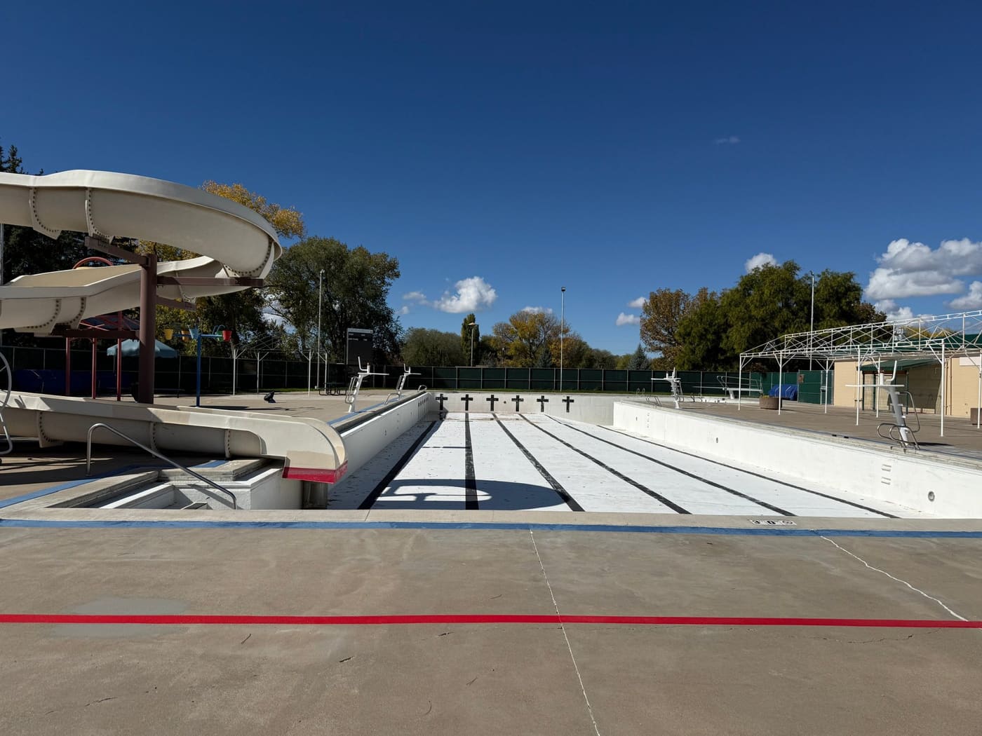 The empty Cortez outdoor pool under a blue sky, with black lane lines on its white floor, lifeguard stands along the deck, and the curving water slide on the left.