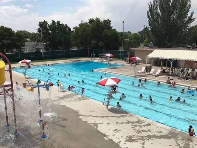 Swimmers fill the Cortez outdoor pool on a sunny summer day, seen from above, with lifeguard stands and umbrellas along the deck and the splash pad's spray features in the foreground.