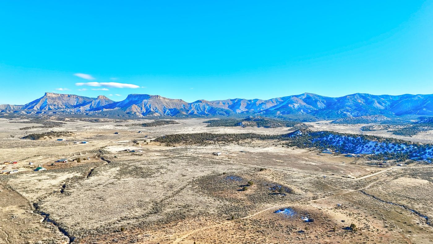 Aerial view over open sagebrush and scattered homes, with snow-dusted mountains and a flat-topped mesa on the horizon under a blue sky.