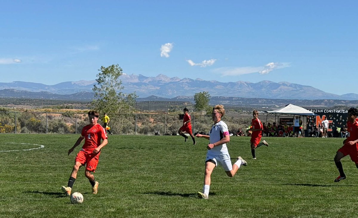 A Montezuma-Cortez player in an orange jersey pushes the ball upfield with a Pagosa Springs player in white running alongside, with mountains on the horizon on a sunny afternoon.