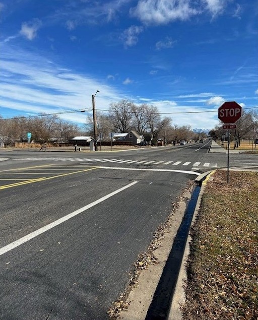 A paved intersection with fresh white striping and a stop sign, where a concrete drainage channel runs along the curb beside a strip of dry grass and fallen leaves.
