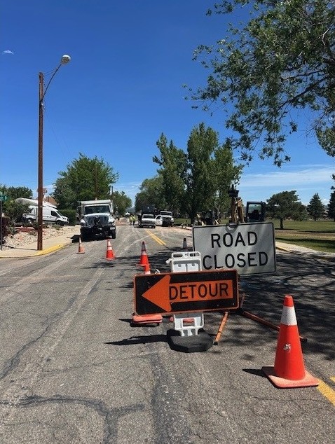 A street closed for work on a sunny day, with Road Closed and Detour signs and orange cones in the lane and work trucks and equipment farther down the block.