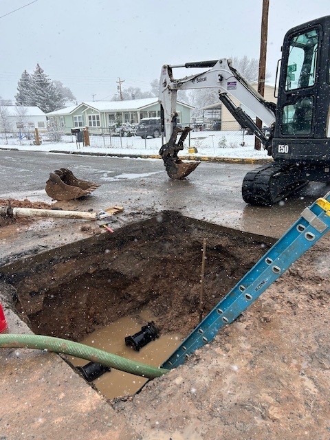 A small excavator beside a muddy, water-filled hole in a snowy street, where crews have exposed a broken water line, with a pump hose and a ladder leading into the hole.