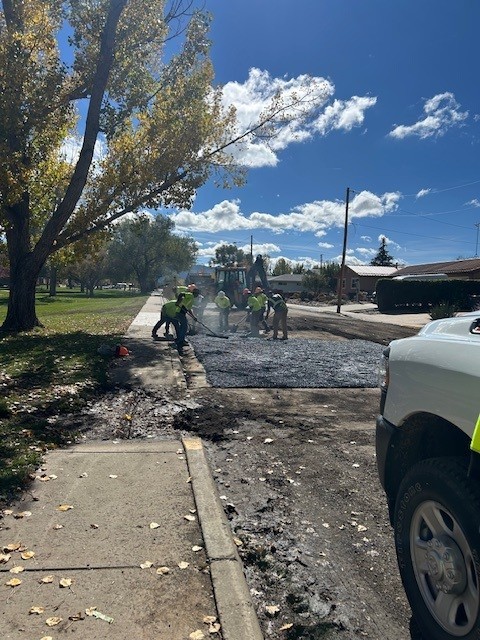 A crew in green safety vests rakes fresh asphalt into a patched section of street beside a park, with a backhoe behind them and a city pickup parked at right.