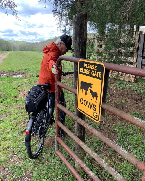 A cyclist in a red jacket opens a metal ranch gate posted with a yellow sign reading Please Close Gate, Cows.