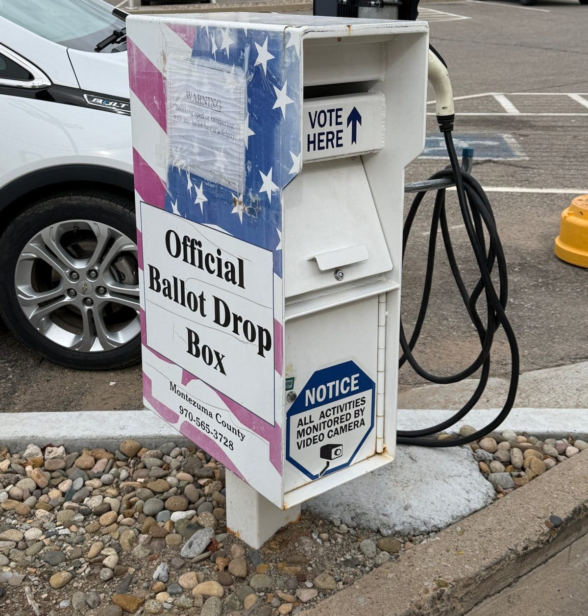 A white Montezuma County ballot drop box decorated with stars and stripes, labeled Official Ballot Drop Box and Vote Here, beside an electric-vehicle charger.